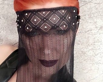 May include: A black lace mask with rhinestones covers the face of a person with bright red hair. The mask has a delicate pattern and is sheer, allowing the person's features to be seen through the fabric.