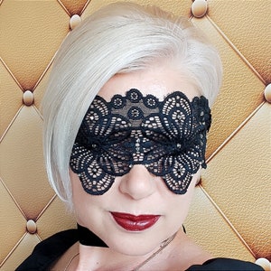 May include: A black lace eye mask with a floral design. The mask is made of delicate lace and features a scalloped edge. The mask is worn over the eyes and is secured with a black ribbon or elastic band. The mask is a dark color.