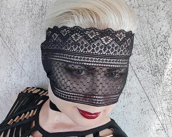 May include: A person wearing a black lace mask over their eyes. The mask has a delicate pattern and covers the entire face. The person is wearing a black top with gold accents.