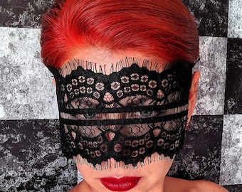 May include: A person with bright red hair wearing a black lace mask over their eyes. The mask has a delicate floral pattern and a scalloped edge. The person is wearing red lipstick and has a neutral expression.