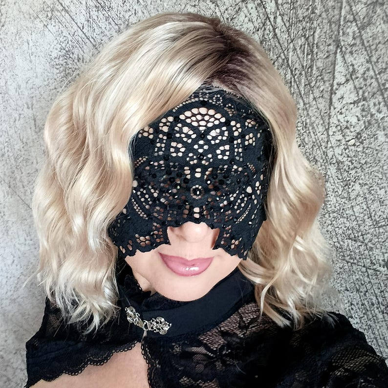 May include: A black lace eye mask with an intricate floral design. The mask covers the eyes and is worn with a black lace top. The person has blonde hair and is wearing a black choker with a decorative clasp.