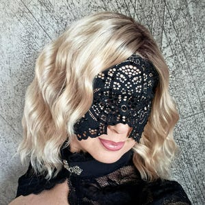 May include: A black lace eye mask with a delicate floral pattern. The mask covers the eyes and part of the nose. The person is wearing a black top with a decorative brooch. The hair is blonde and wavy.