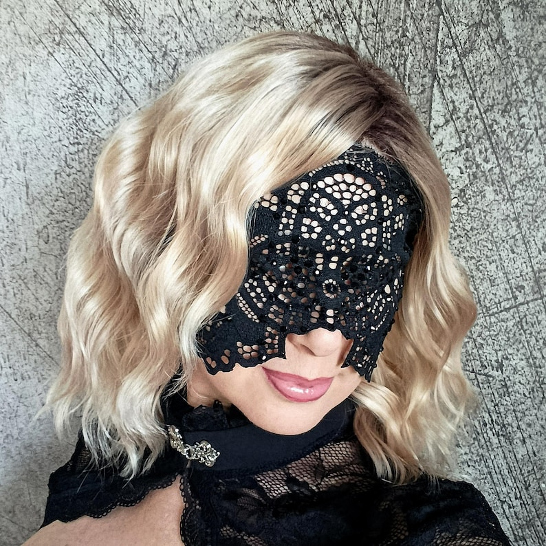 May include: A black lace eye mask with a delicate floral pattern. The mask covers the eyes and part of the nose. The person is wearing a black top with a decorative brooch. The hair is blonde and wavy.