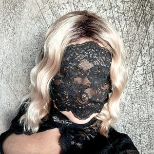 May include: A black lace face mask adorned with gold-coloured embellishments. The mask covers the eyes and nose, with a scalloped edge. The person is wearing a black top and has wavy blonde hair.