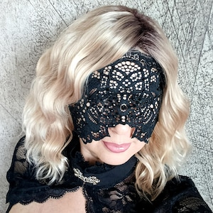 May include: A black lace eye mask with an intricate floral design. The mask covers the eyes and nose, with openings for the mouth. The person is wearing a black lace top and has wavy blonde hair.