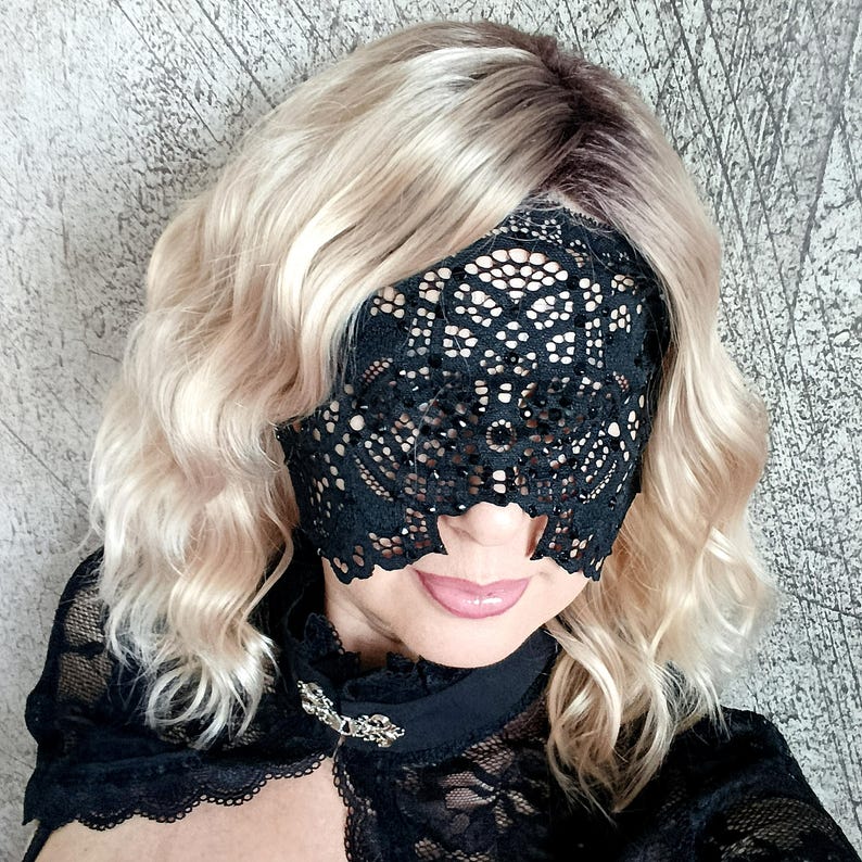 May include: A black lace eye mask with an intricate floral design. The mask covers the eyes and nose, with openings for the mouth. The person is wearing a black lace top and has wavy blonde hair.
