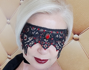 May include: A black lace eye mask with red rhinestones and a single larger red rhinestone in the center. The mask is worn by a person with short, platinum blonde hair.
