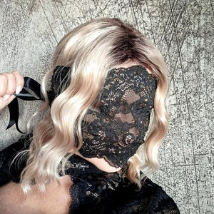 May include: A black lace mask with a floral pattern covers the face. The mask is embellished with small, sparkling accents. A black satin ribbon is held to the side. The person has wavy blonde hair.