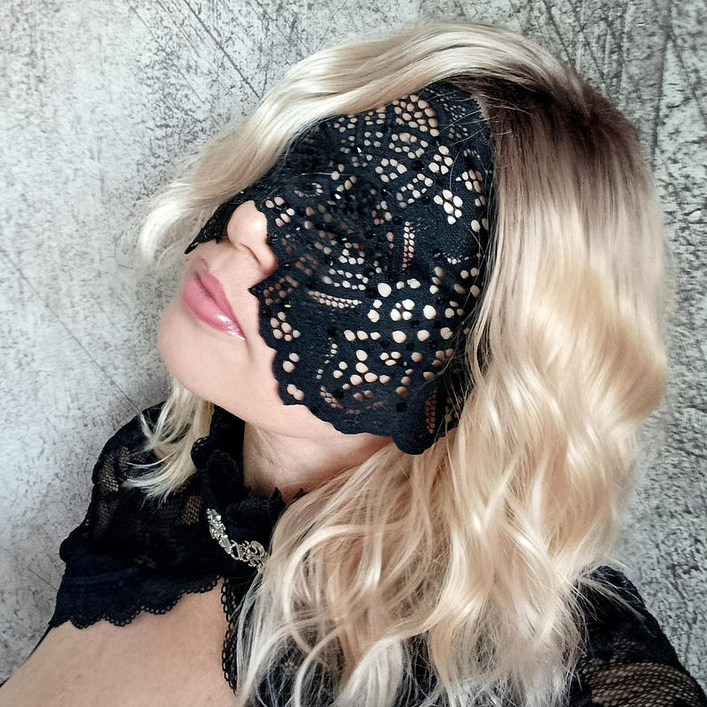 May include: Black lace eye mask with a floral pattern. The mask covers the eyes and is worn over the face. The person in the image has blonde hair and is wearing a black top.
