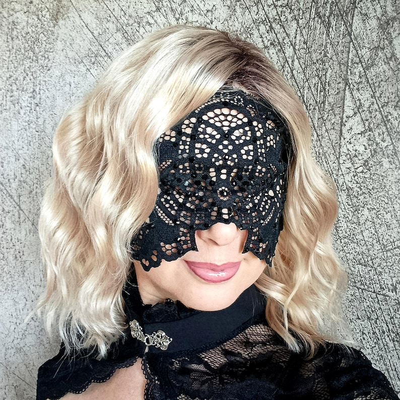 May include: A black lace eye mask with an intricate floral design. The mask covers the eyes and part of the nose. The person is wearing a black top with a decorative clasp.