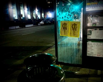 Puede incluir: Una vista nocturna de una parada de autobús con un contenedor de basura en primer plano. La parada de autobús tiene una ventana de vidrio con grafitis y dos carteles amarillos con un diseño psicodélico. La parada de autobús también tiene un letrero que dice "City of Oakland".