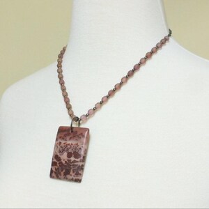 May include: A pink and brown stone pendant necklace with a rectangular pendant. The pendant has a marbled pattern with shades of pink, brown, and white.