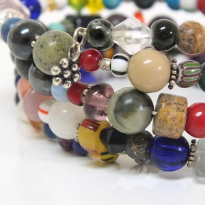May include: Multi-strand bracelet featuring a variety of colorful beads, including red, blue, green, brown, and white. The bracelet is made with natural stones, glass beads, and metal accents.