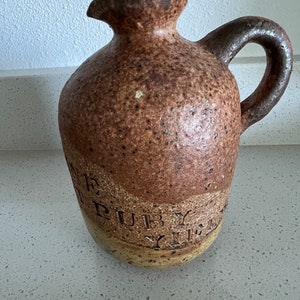 May include: A brown ceramic jug with a cork stopper and a handle. The jug has a speckled glaze and the words "The Pub" are visible on the side.