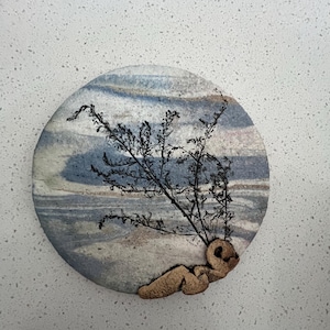 May include: A round ceramic wall hanging with a blue and white marbled design. A brown, textured, 3D sculpture of a person is attached to the bottom of the circle. The sculpture is lying on its side with its head resting on its hand.