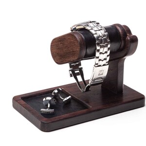 May include: A wooden watch and cufflinks stand with a black leather watch holder and a black leather cufflinks tray. The stand is dark brown and has a smooth finish.