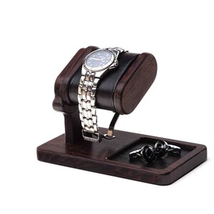 May include: A dark brown wooden watch stand with a black leather cushion for a watch. The stand has a small compartment for cufflinks or other jewelry. The watch stand is on a white background.