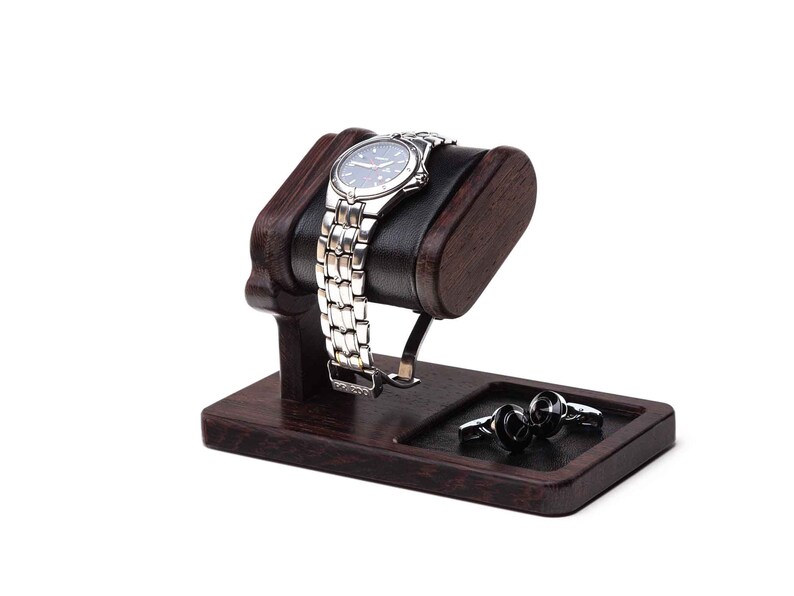 May include: A dark brown wooden watch stand with a black leather cushion for a watch. The stand has a small compartment for cufflinks or other jewelry. The watch stand is on a white background.