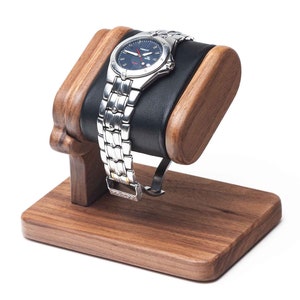 May include: A wooden watch stand with a black leather cushion to hold a watch. The stand is made of dark wood and has a simple design.