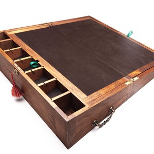 Op de afbeelding: Een houten kist met een donkerbruin interieur en een scharnierend deksel. De kist heeft verschillende compartimenten en een handvat. Een rode kwast en een groen lint zijn aan de kist bevestigd.