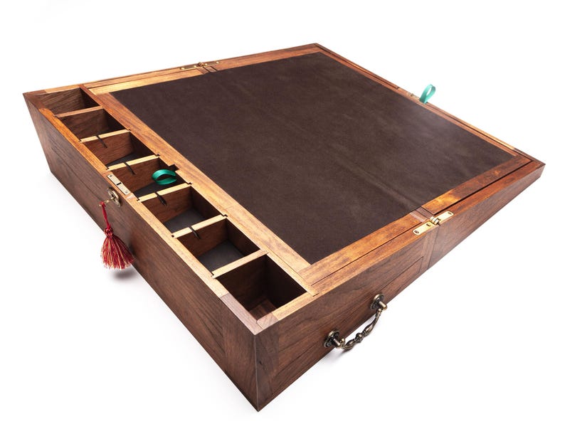 Op de afbeelding: Een houten kist met een donkerbruin interieur en een scharnierend deksel. De kist heeft verschillende compartimenten en een handvat. Een rode kwast en een groen lint zijn aan de kist bevestigd.