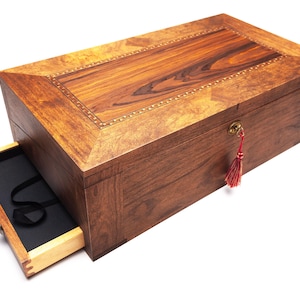 Op de afbeelding: Een rechthoekige houten kist met een gedeeltelijk open lade, die een zwart interieur onthult. De kist is gemaakt van donkerbruin hout met een lichtere houten inleg op het deksel. Een rode kwast hangt aan het sleutelgat.
