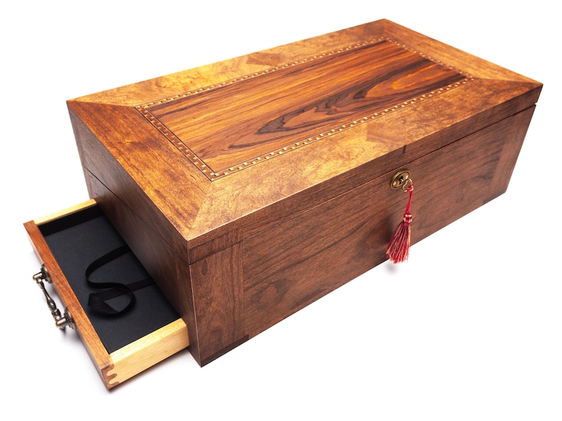Op de afbeelding: Een rechthoekige houten kist met een gedeeltelijk open lade, die een zwart interieur onthult. De kist is gemaakt van donkerbruin hout met een lichtere houten inleg op het deksel. Een rode kwast hangt aan het sleutelgat.