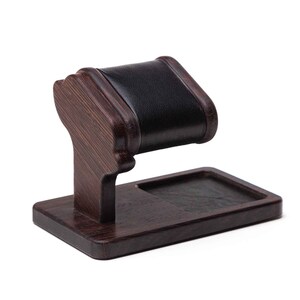 May include: A dark brown wooden watch stand with a black leather cushion for holding a watch. The stand has a small rectangular tray for holding other items.