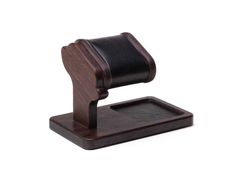 May include: A dark brown wooden watch stand with a black leather cushion for holding a watch. The stand has a small rectangular tray for holding other items.