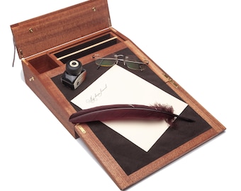Op de afbeelding: Een houten bureau met een scharnierend deksel, open om een ​​zwart vilten schrijf oppervlak te onthullen. Een pen, inktpot en een bril liggen op het oppervlak. Het bureau heeft een klein compartiment voor opslag.