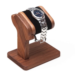 May include: A wooden watch stand with a black padded arm to hold a watch. The stand is made of dark wood and has a square base.