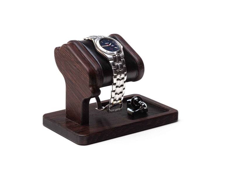 May include: A dark brown wooden watch stand with a black leather cushion to hold a watch. A pair of cufflinks are on the stand's tray.
