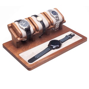 Puede incluir: Un expositor de relojes de madera con un forro de terciopelo beige claro. El expositor contiene cuatro relojes de pulsera con correas de cuero y metal, y un reloj inteligente negro con una correa de silicona. La madera tiene un acabado marrón cálido.