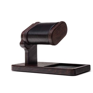 May include: A dark brown wooden watch stand with a black leather cushion. The stand has a rectangular base and a curved arm with a cushion for holding a watch.
