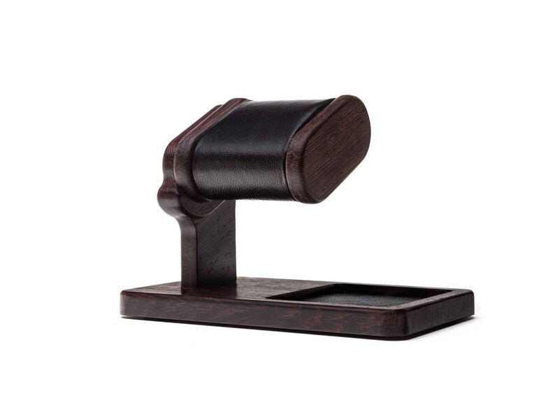 May include: A dark brown wooden watch stand with a black leather cushion. The stand has a rectangular base and a curved arm with a cushion for holding a watch.