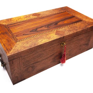Op de afbeelding: Een rechthoekige houten kist met een scharnierend deksel. De kist is gemaakt van donkerbruin hout met een lichtere houten inleg op het deksel. Een decoratief sleutelgat en een rode kwast voegen details toe. De kist heeft metalen hardware.