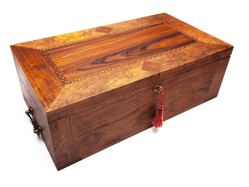 Op de afbeelding: Een rechthoekige houten kist met een scharnierend deksel. De kist is gemaakt van donkerbruin hout met een lichtere houten inleg op het deksel. Een decoratief sleutelgat en een rode kwast voegen details toe. De kist heeft metalen hardware.