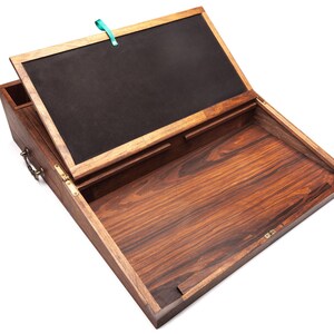 Op de afbeelding: Een houten schrijfkist met een scharnierend deksel, open om een donker interieur te onthullen. De kist is gemaakt van donker hout met een zichtbaar nerfpatroon. Een kleine penhouder is aan de zijkant bevestigd. Een turquoise lint is aan het deksel bevestigd.