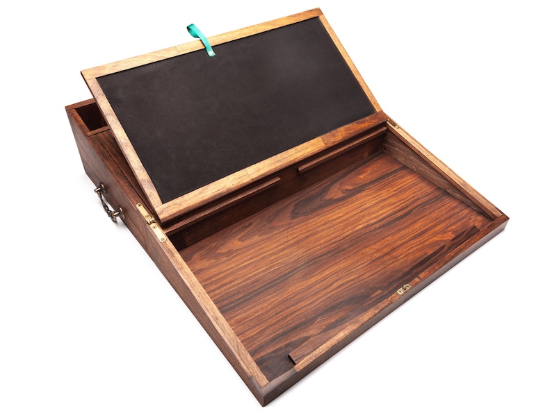 Op de afbeelding: Een houten schrijfkist met een scharnierend deksel, open om een donker interieur te onthullen. De kist is gemaakt van donker hout met een zichtbaar nerfpatroon. Een kleine penhouder is aan de zijkant bevestigd. Een turquoise lint is aan het deksel bevestigd.