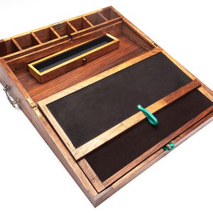 Op de afbeelding: Een houten bureau-organizer met meerdere compartimenten en een scharnierend deksel. De organizer heeft een donkere houten afwerking met een zwart interieur en een kleine lade. Een turquoise lint is aan het deksel bevestigd.