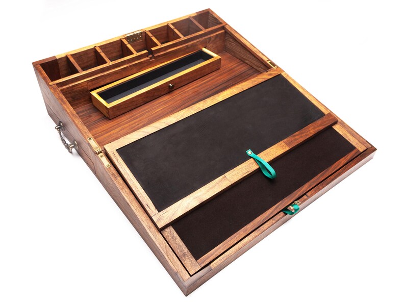 Op de afbeelding: Een houten bureau-organizer met meerdere compartimenten en een scharnierend deksel. De organizer heeft een donkere houten afwerking met een zwart interieur en een kleine lade. Een turquoise lint is aan het deksel bevestigd.