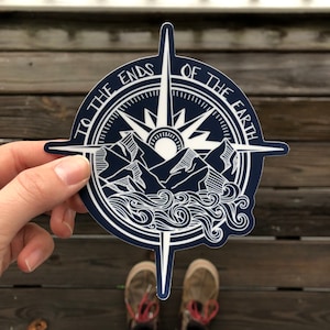 May include: A navy blue compass sticker with white details. The compass features a sun, mountains, and waves. The text around the compass reads "To the Ends of the Earth".