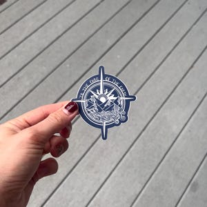 Può includere: Un adesivo di bussola blu con un design bianco. La bussola presenta una catena montuosa, un sole e il testo "To the Ends of the Earth".