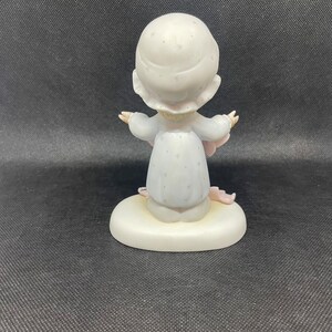 May include: A porcelain figurine of a girl wearing a white dress with a pink floral pattern and a white hat with a floral pattern. She is holding a pink blanket and is sitting on a white base.