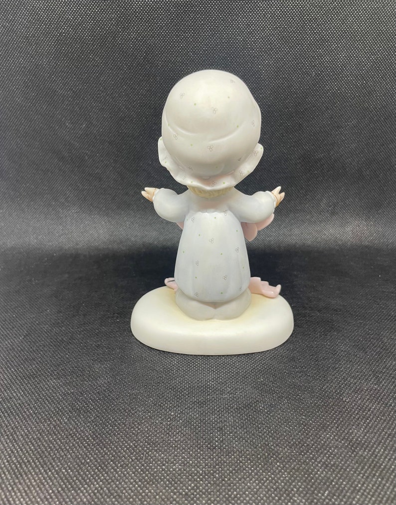 May include: A porcelain figurine of a girl wearing a white dress with a pink floral pattern and a white hat with a floral pattern. She is holding a pink blanket and is sitting on a white base.