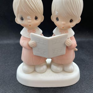 May include: A ceramic figurine featuring two figures in pink robes and white collars, holding a sheet of music. The figures have light-colored hair and are standing on a white base. The figurine has a classic, vintage aesthetic.