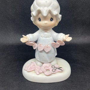May include: A porcelain figurine of a girl in a nightgown holding a string of pink hearts. She is standing on a base with a pair of scissors and a pink ribbon.