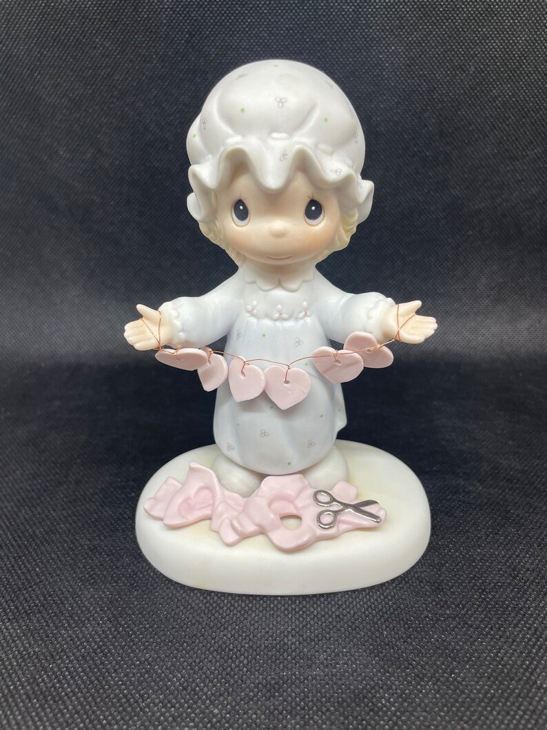 May include: A porcelain figurine of a girl in a nightgown holding a string of pink hearts. She is standing on a base with a pair of scissors and a pink ribbon.