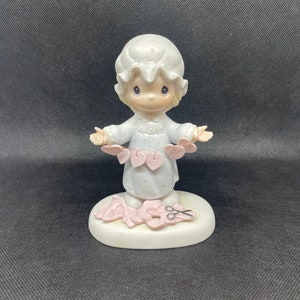 May include: A porcelain figurine of a girl wearing a white nightgown and holding a string of pink hearts. She is standing on a base with a pair of scissors and more pink hearts.