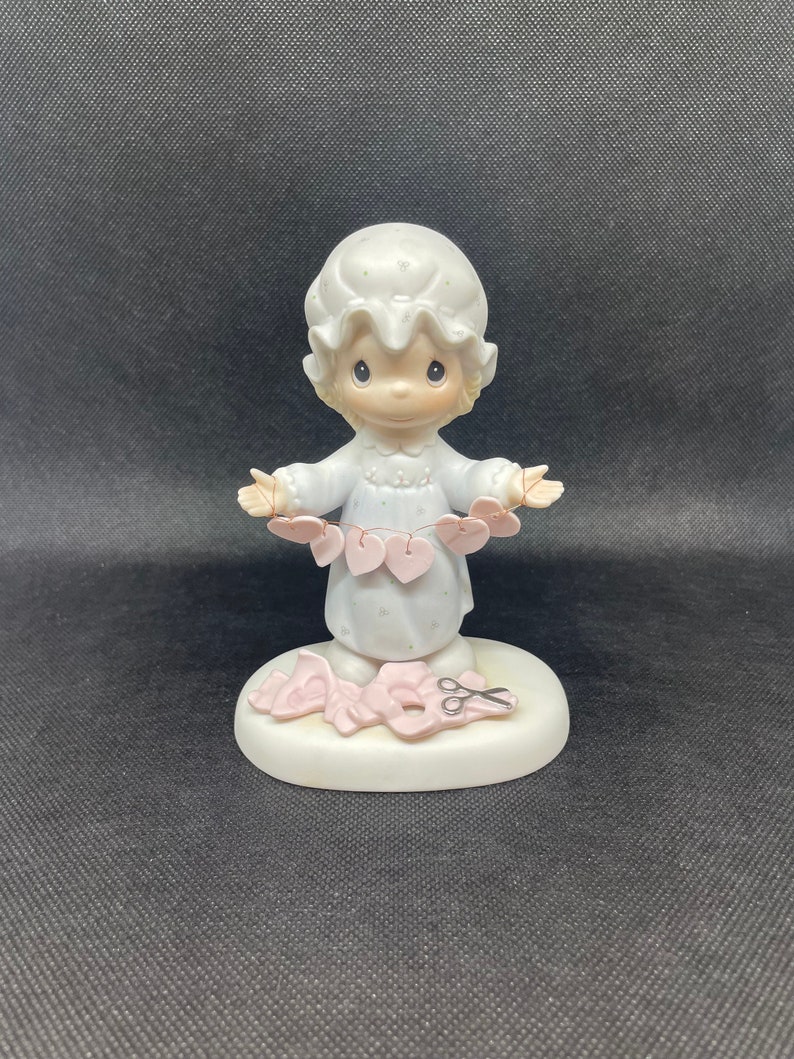May include: A porcelain figurine of a girl wearing a white nightgown and holding a string of pink hearts. She is standing on a base with a pair of scissors and more pink hearts.